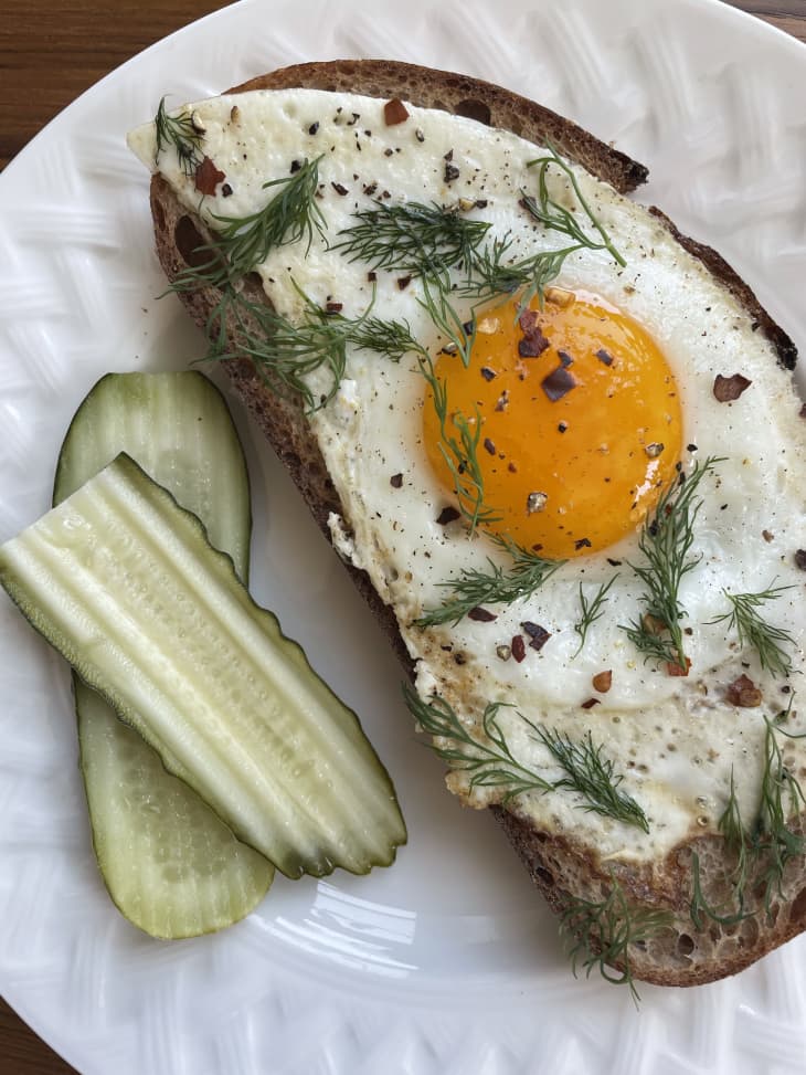 Plated pickle brine fried egg on toasted bread, garnished with dill and red pepper flakes. Pickles on the side.