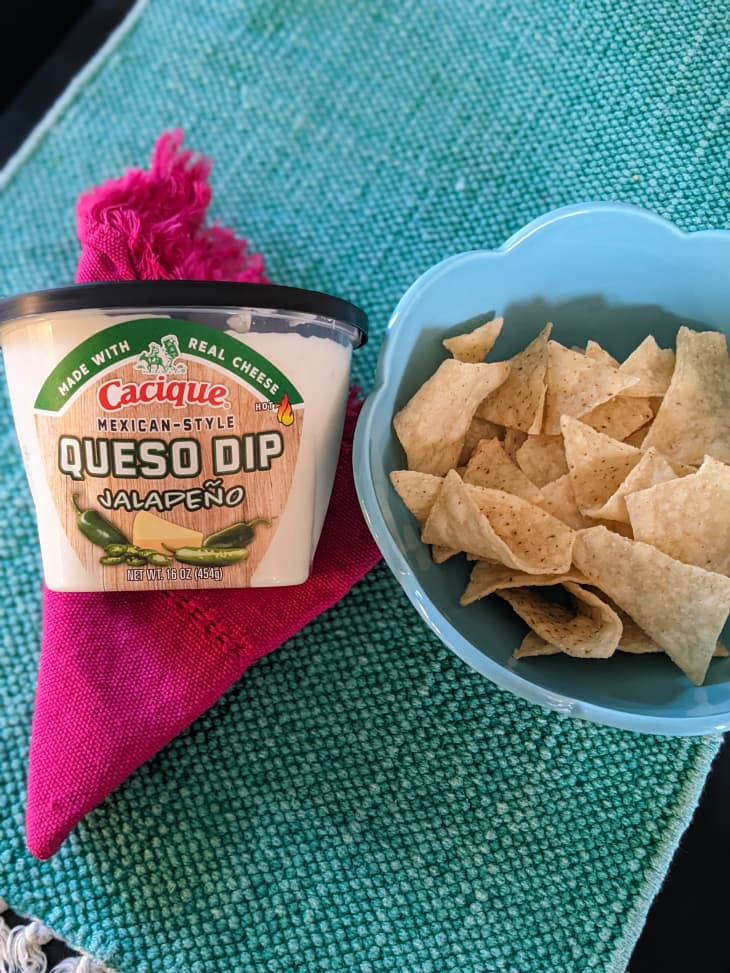 Cacique queso dip in package beside a bowl of tortilla chips.