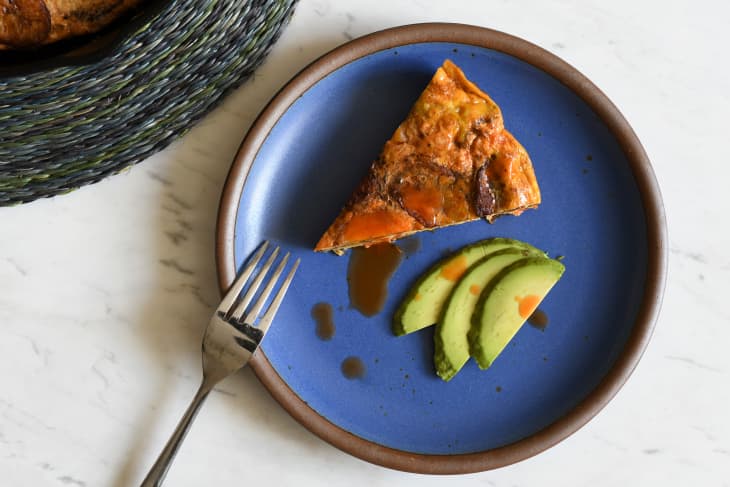 Plated slice of plantain frittata with side of avocado and drizzle of hot sauce on top.