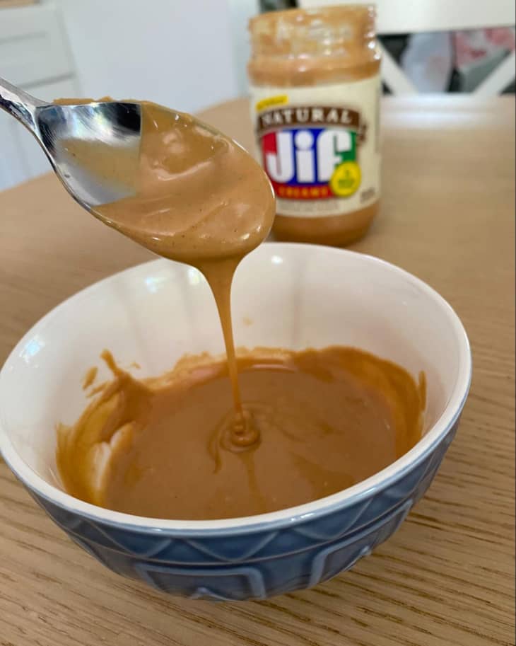 Melted peanut butter in a bowl on a table. Someone is scooping it with a spoon. Jar of Jif Peanut Butter in the background.