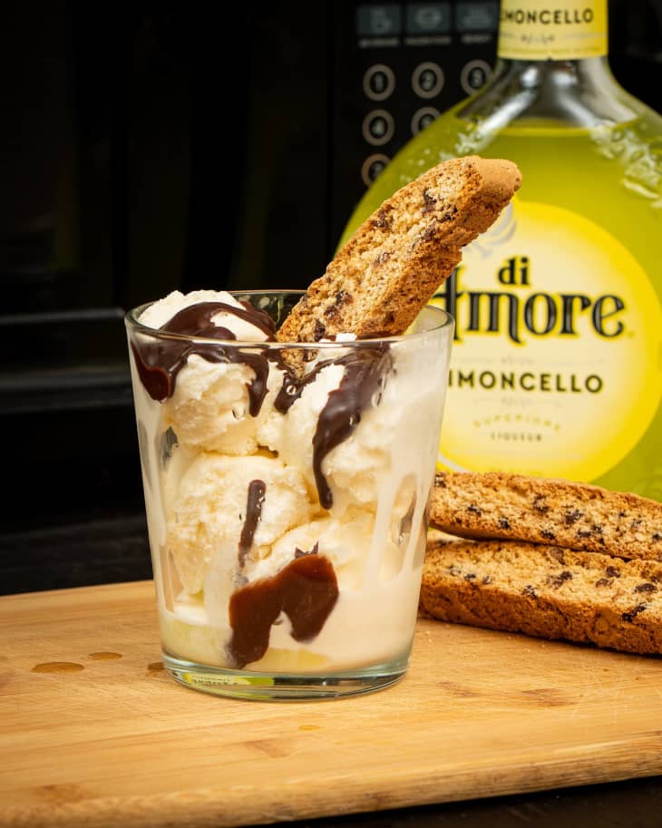 glass with ice cream, biscotti, and chocolate sauce on cutting board with biscotti and bottle of limoncello in the background.