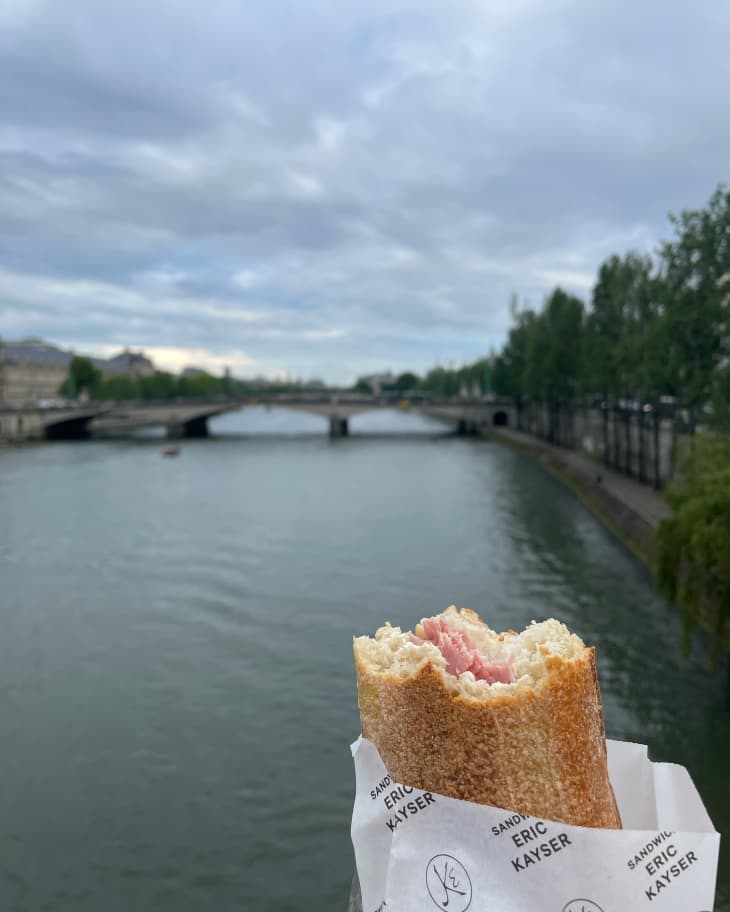 someone holding a paper wrapped ham and butter sandwich on baguette in front of the Seine River in Paris