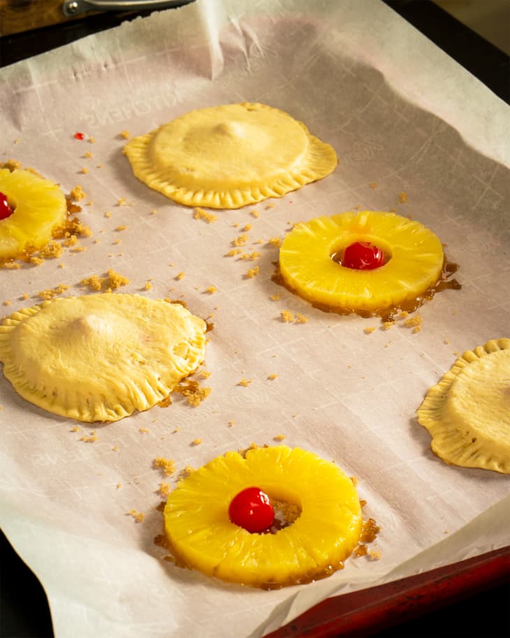 Wax paper lined tray topped with cherries inside pineapple rings before baking. Some cherry filled pineapple rings have been wrapped in pastry dough before baking.