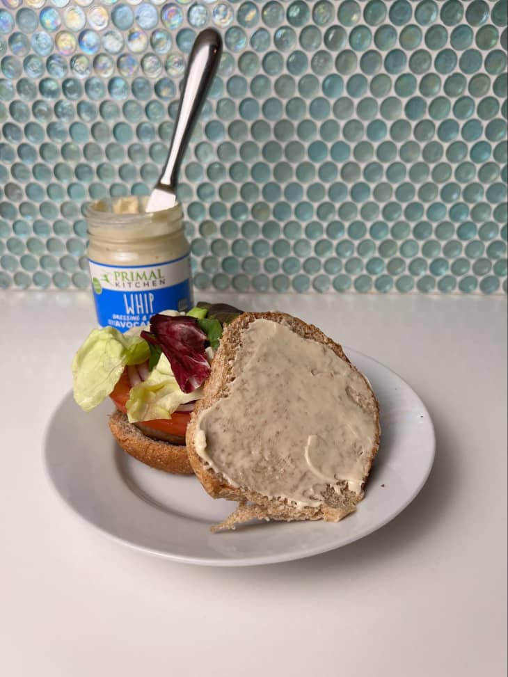 Sandwich on plate with top piece of bread opened showing condiment on top. Jar of Primal Kitchen's whip dressing and spread in the background.