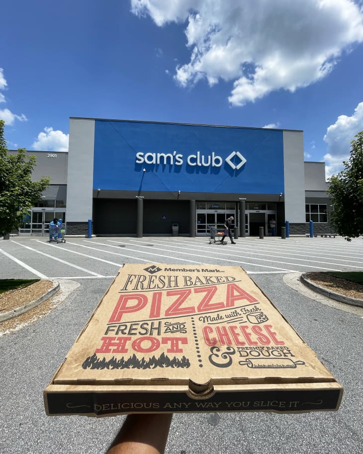 someone holding up a pizza box in front of a sam's club store