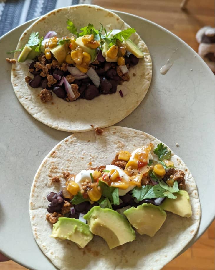 grey plate, wood table, tacos, avocado, cilantro, black beans, drizzle of white sauce, meat pieces