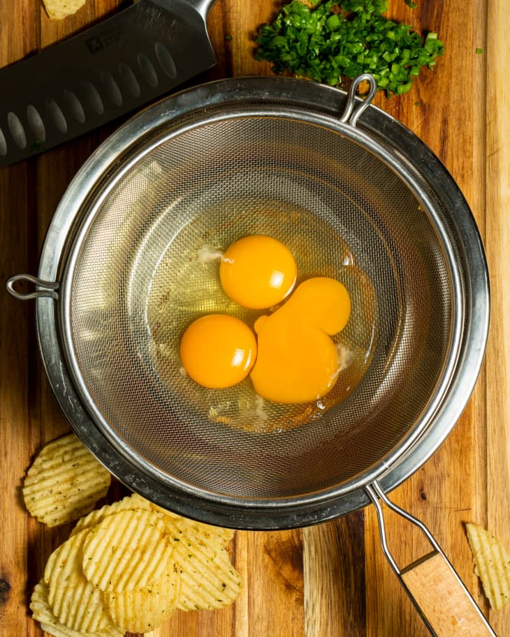 Egg yolks and whites in strainer placed in larger metal bowl with chopped parsley and potato chips on cutting board.