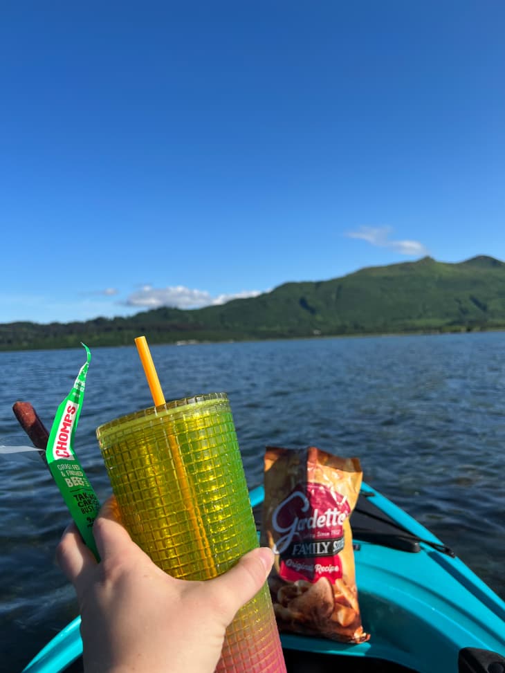 Someone in kayak holding reusable water cup and beef jerky. Bag of snack mix sits on front of kayak.