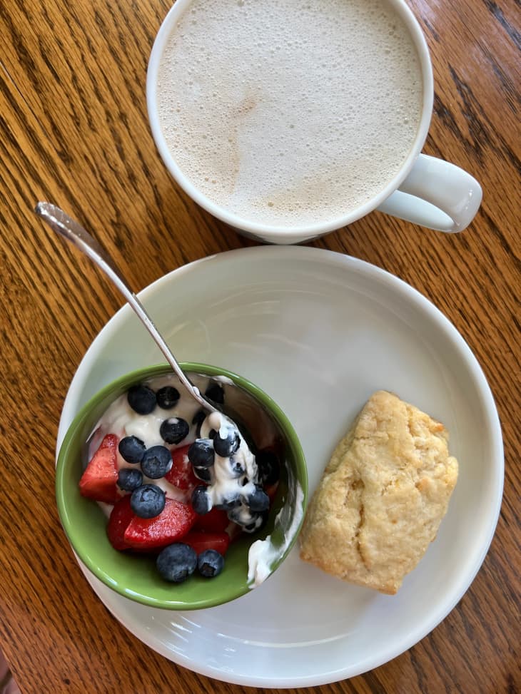 Scone on plate next to bowl of yogurt with strawberries and blueberries. Latte in mug above plate.