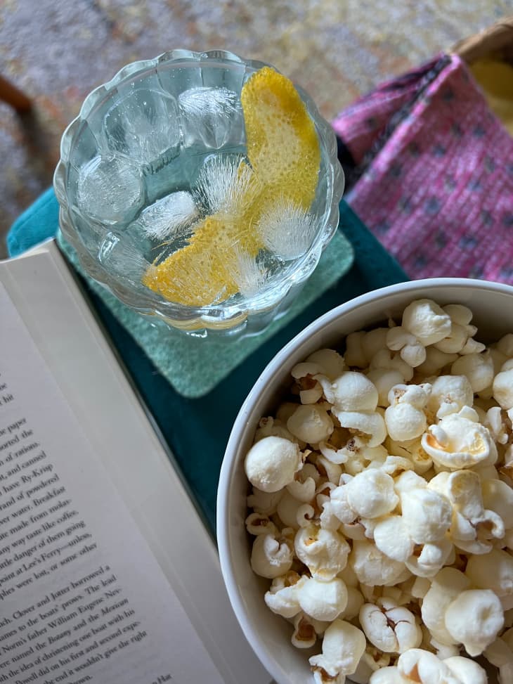 Book on the side of cocktail with lemon peel and bowl of popcorn.