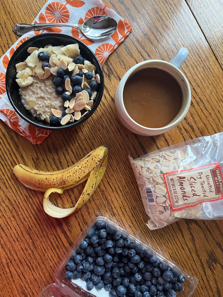 Bowl of oatmeal topped with bananas, blueberries and almonds with mug of coffee on the side. Banana peel, package of blueberries and almonds surround.