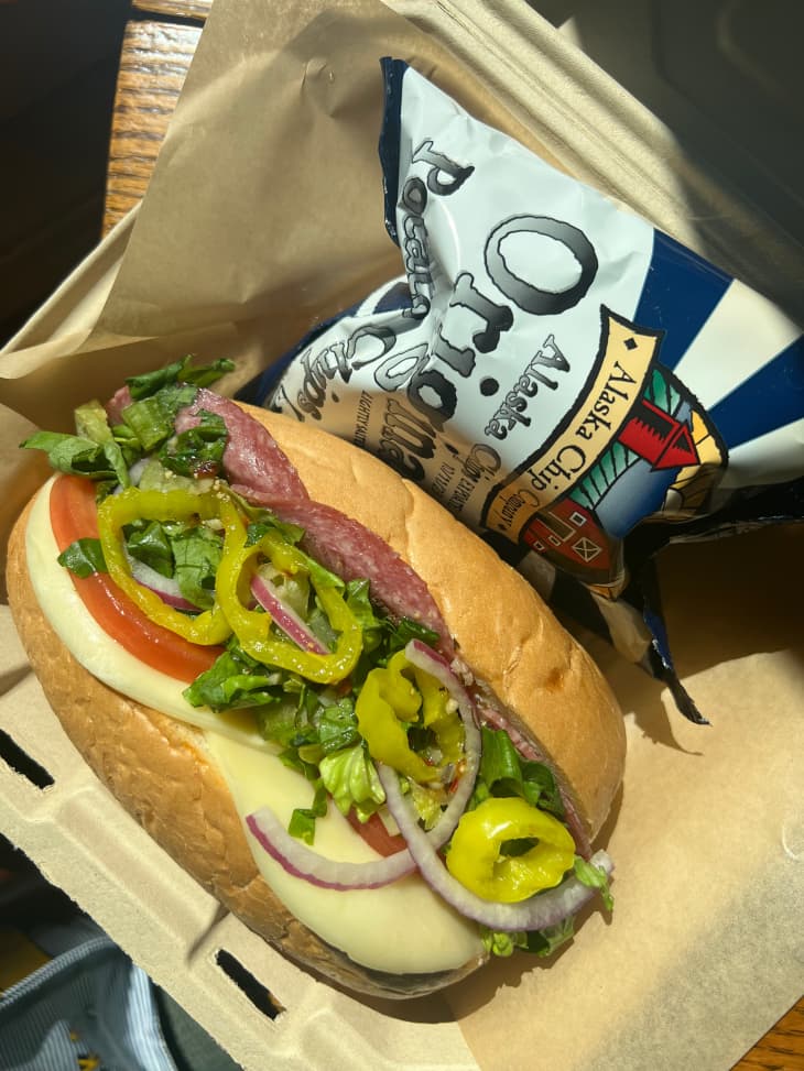 Sub sandwich with meat and cheese topped with pickled peppers, onions and greens. Small bag of potato chips on the side.