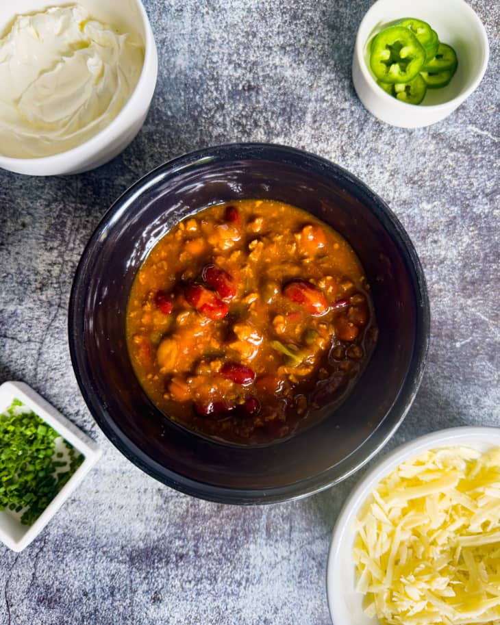 bowl of red chili with cheese, sour cream, chives, and jalepenos in small serving bowls scattered around