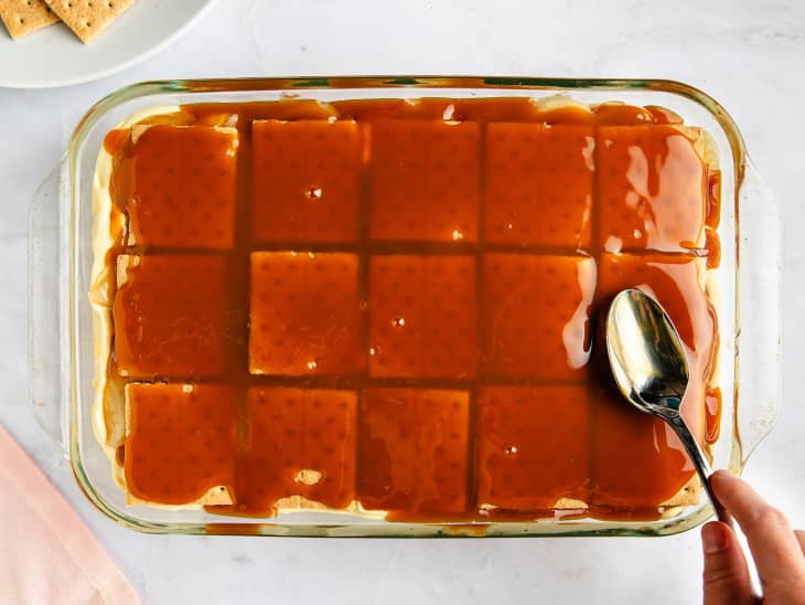 Someone smoothing caramel on top of graham cracker layer to make caramel apple eclair cake.