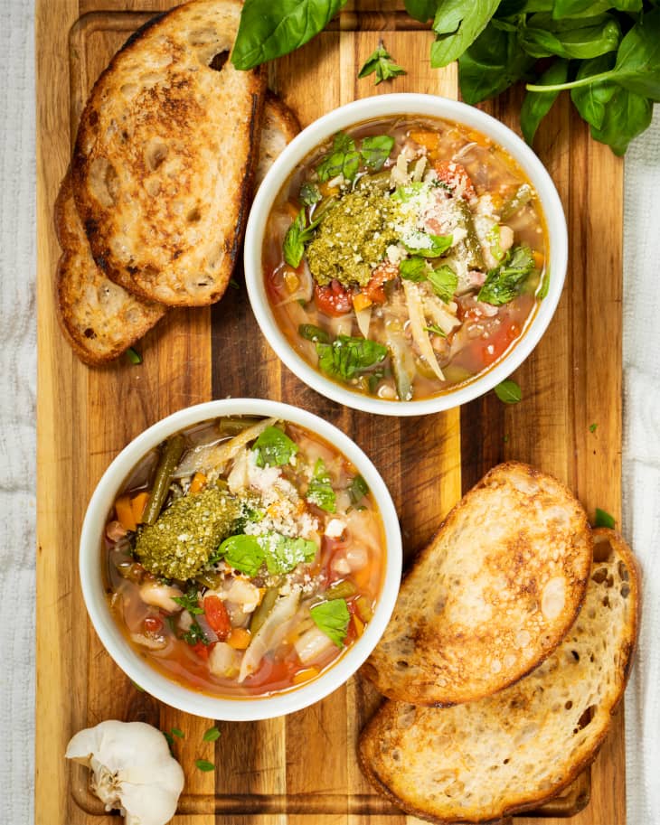 Two bowls of minestrone soup on wooden board with slices of bread on the side.