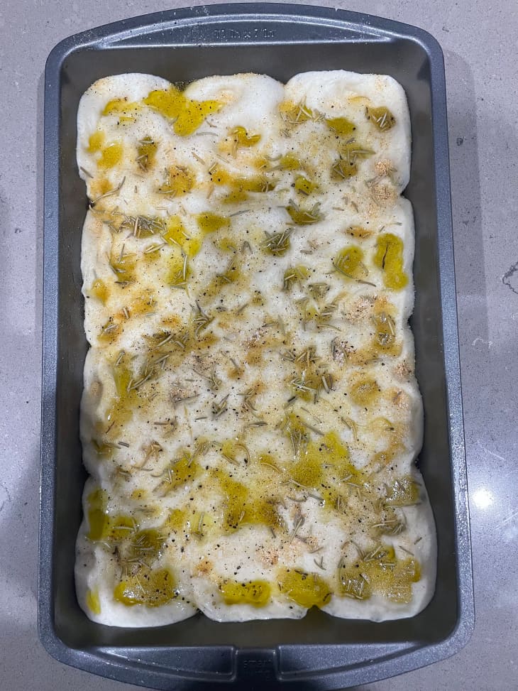 Focaccia dough topped with olive oil and herbs in baking sheet.