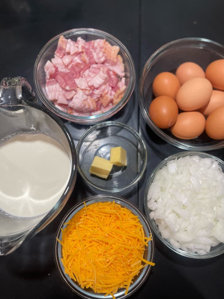 Mise en place of ingredients for cowboy quiche from clockwise top right: eggs, diced onion, cheddar cheese, milk, bacon and butter.