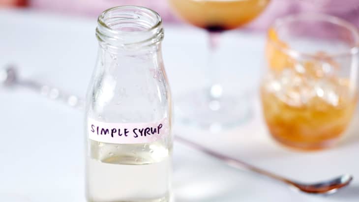 Clear bottle of simple syrup with a handmade label sitting on a table. There are drinks and cocktail stirring spoon in the background