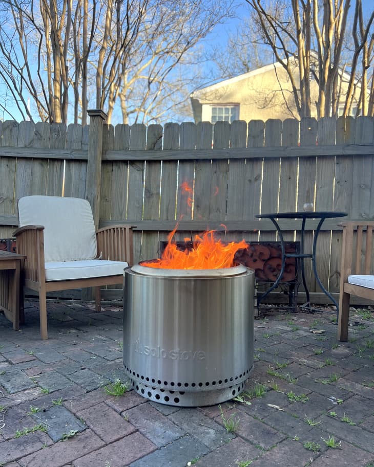 Solo Stove Bonfire burning on back brick patio. Chairs and a table. Fence behind with trees