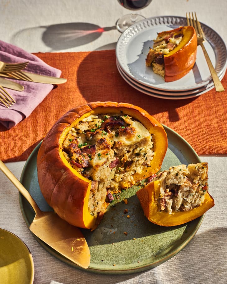 Pumpkin stuffed with bread cubes, kale, bacon on sage green plate. Slice has been served on smaller white plate. There is gold flatware and servingware, and a cloth napkin. A glass of red wine is in the upper right
