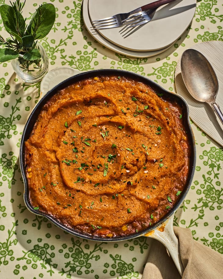 sweet potato and butternut squash shepherd's pie in a skillet on a thanksgiving table. There is a spoon next to it, and a small bouquet of sage and rosemary in a glass vase. some extra plates and forks are in the back