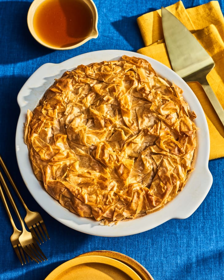 Overhead photo of a whole apple pear pie. The crust (phyllo) on top is very intricate and folded. There is a bright blue tablecloth, bright gold napkin, gold servingware and plates in shot