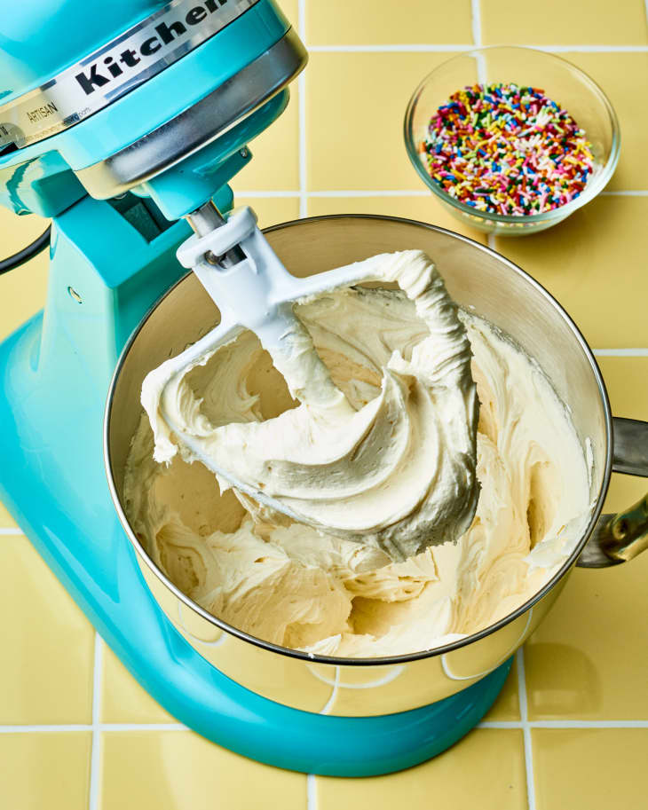 aqua blue stand mixer mixing buttercream frosting on yellow tile countertop. Bowl of colored sprinkles in the background