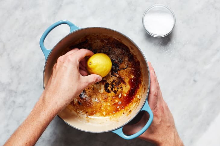 Someone squeezing lemon into burnt Dutch oven with small bowl of salt off to the side.