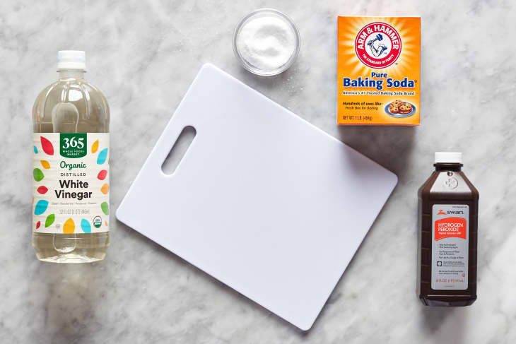 Plastic cutting board laid out with cleaning ingredients surrounded; left to right: White vinegar, kosher salt, baking soda, hydrogen peroxide.