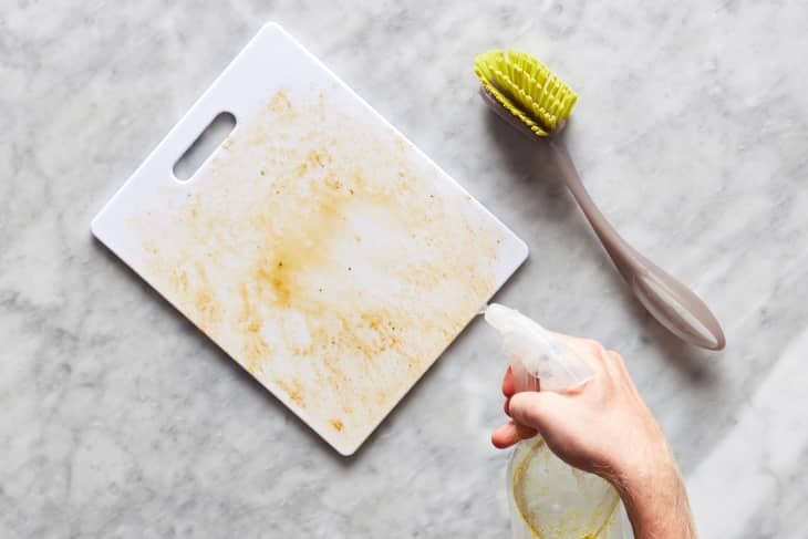 Lemon essential oil being sprayed on soiled cutting board with scrub brush on the side.
