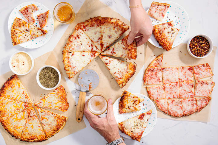 A bunch of baked frozen pizzas on a counter cut up with different toppings in small bowls and two hands reaching in to grab a slice
