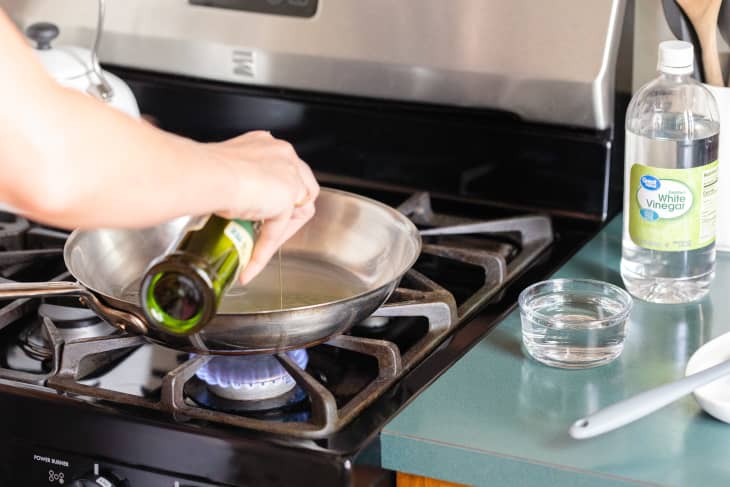 Pouring olive oil into a stainless steel skillet, a bowl of white vinegar and bottle of vinegar next to the stove