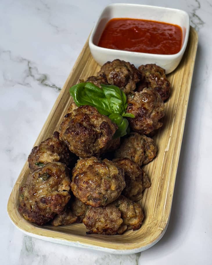 15 small meatballs sitting on a rectangular plate with red sauce in a small square bowl on the plate, with three basil leaves on top