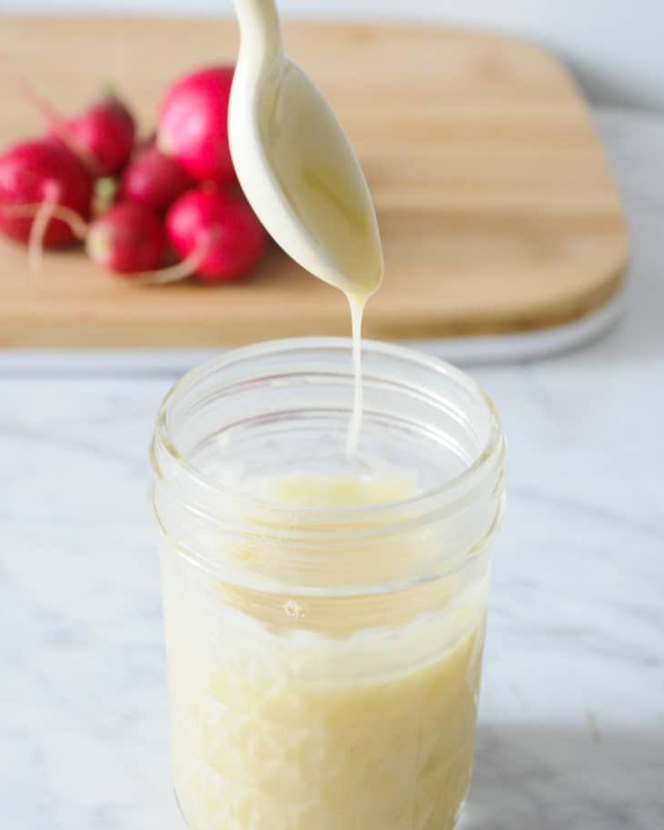 Buerre Blanc sauce in a small  jar, with a spoon held up above the jar, with sauce dripping from it, and radishes in the background