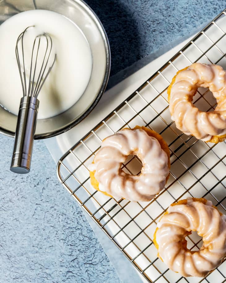 Three donuts with white glaze on a cooling rack, with a whisk in a bowl of glaze next to it