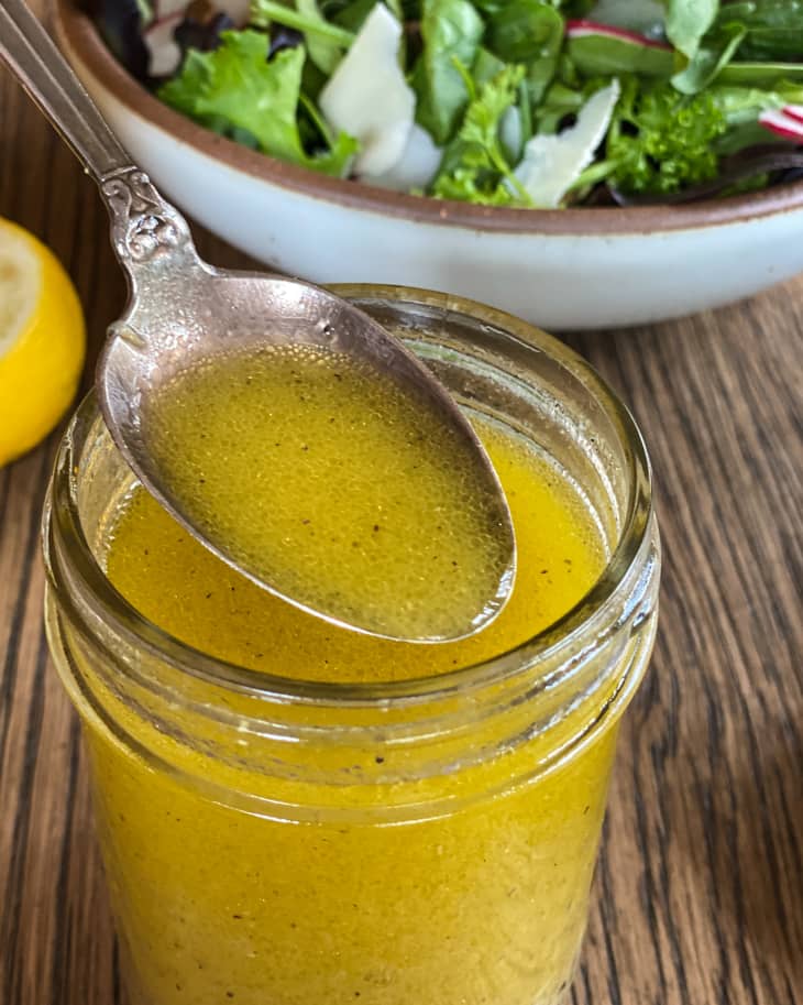 Lemon vinaigrette in a ball jar with a spoonful of the dressing being lifted. There is a green salad in the background.