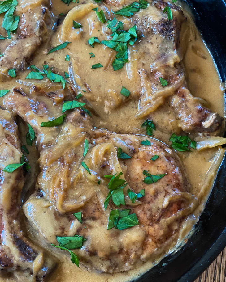 A closeup of Smothered Pork Chops in a black skillet with brown gravy and and green garnish on top.