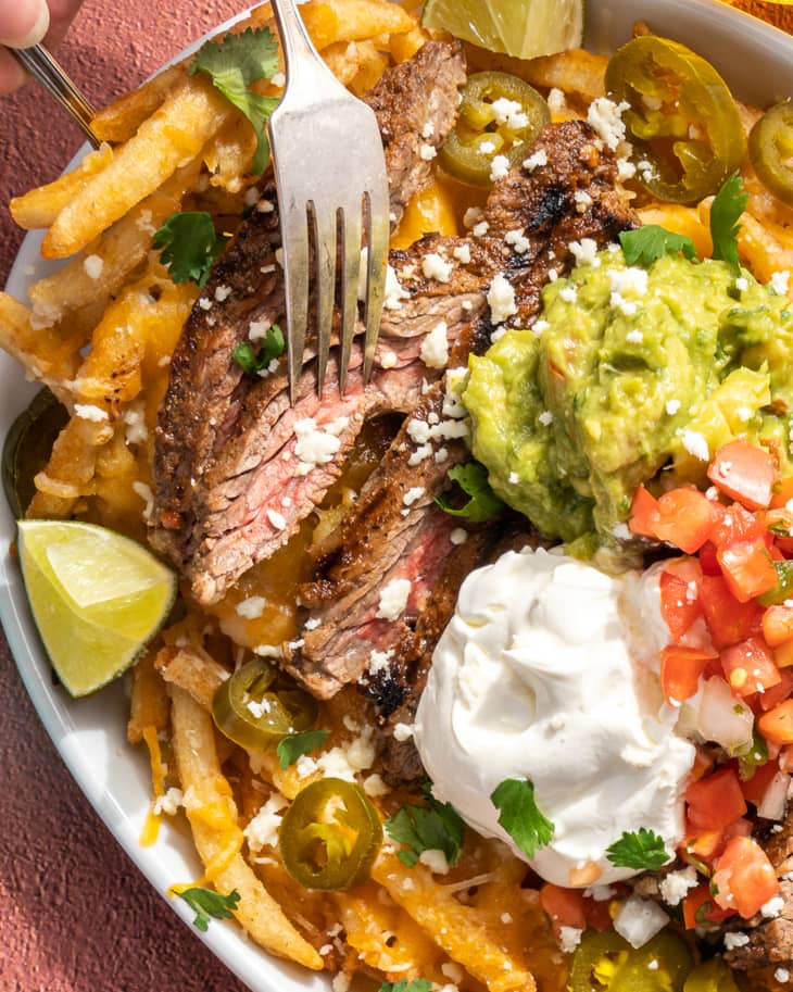 french Fries with Carne Asada beef. guacamole, salsa, sour cream, lime wedges and crumbled white cheese on top, surrounded by plates with the aforementioned ingredients.