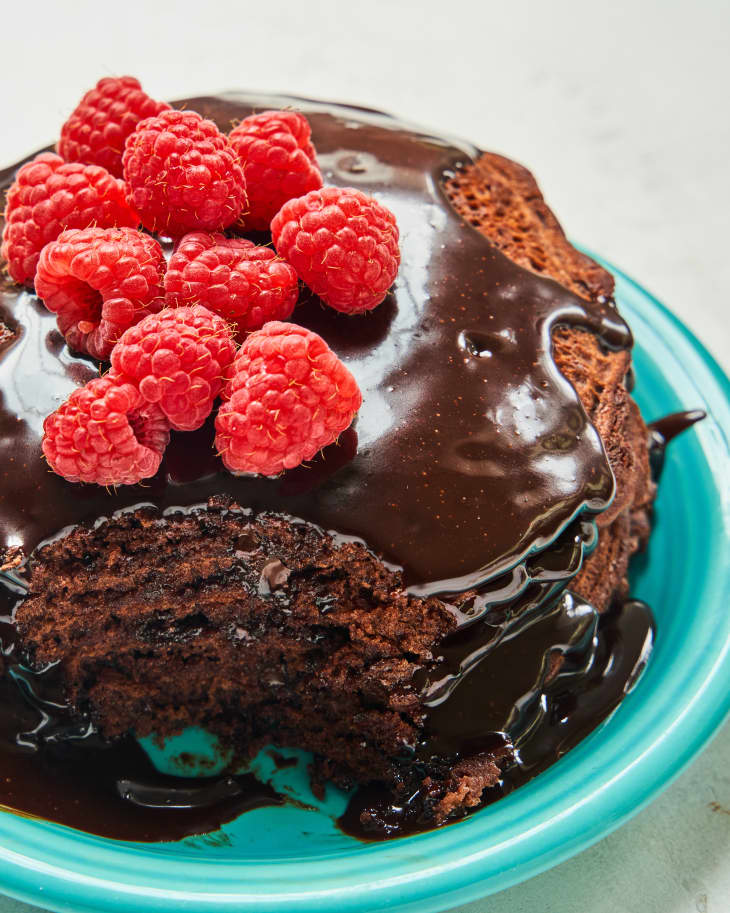 a stack of chocolate pancakes on a plate with chocolate sauce and raspberries on top, with a piece taken out of the front.