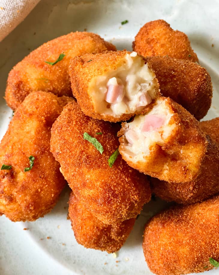 A pile of croquetas on a white plate with a green garnish, with one Croqueta broken in half on top to show the ham and cheese inside.