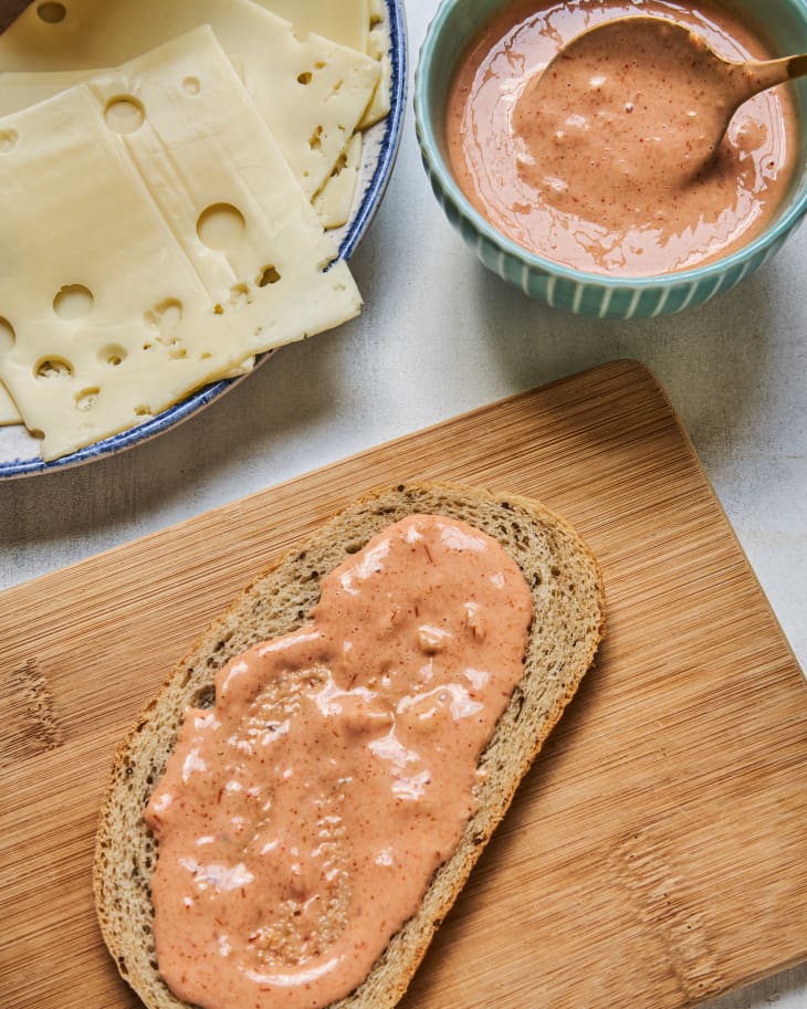 Russian dressing spread on bread, on a wooden platter with a plate with swiss cheese slices and a blue ramekin with russian dressing next to it.