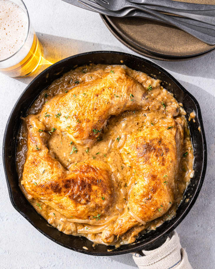 smothered chicken in a pan with a glass of beer and a plate with utensils next to it.