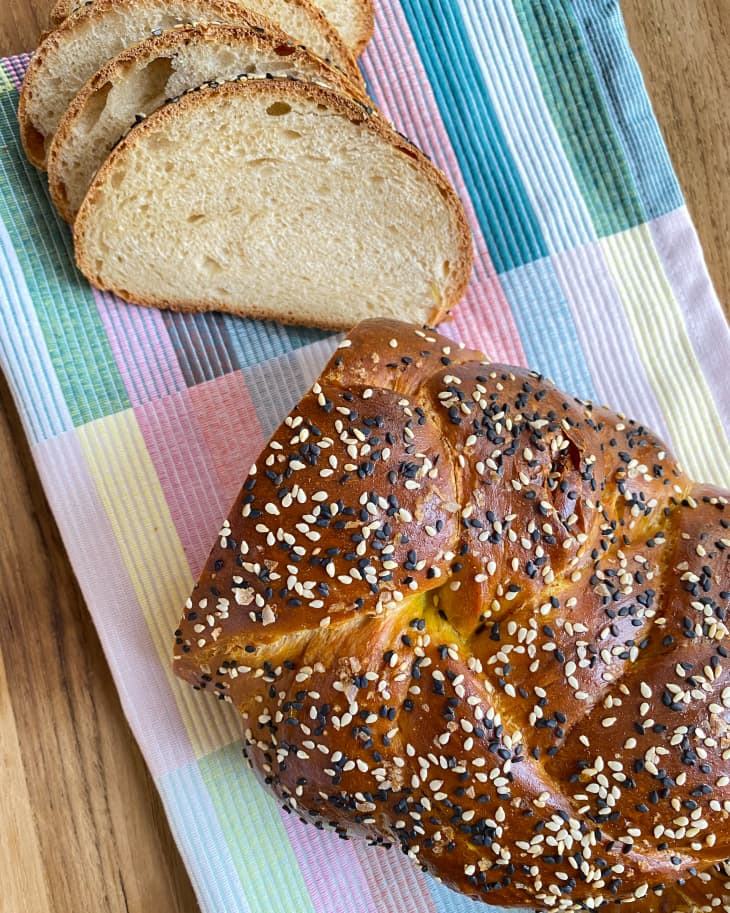 Sourdough Challah (a rich, traditional Jewish bread) with poppy and sesame seeds on top. Half of the loaf is sliced, and it sits atop a pastel rainbow napkin.