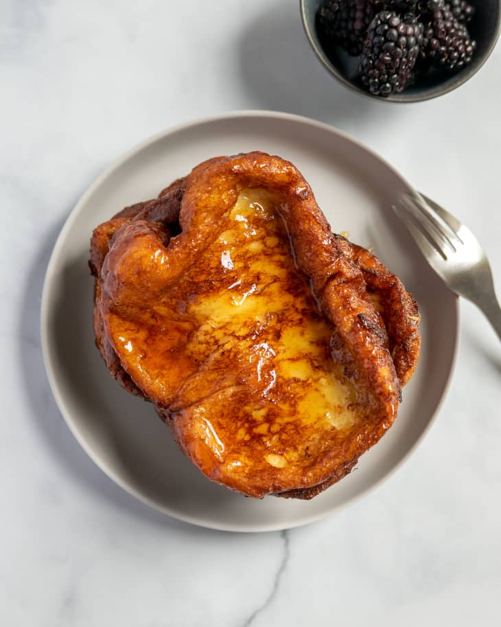 torrijas (french toast)  stacked on a light  gray, circular plate, with a silver fork, and a small bowl of blackberries on the side.