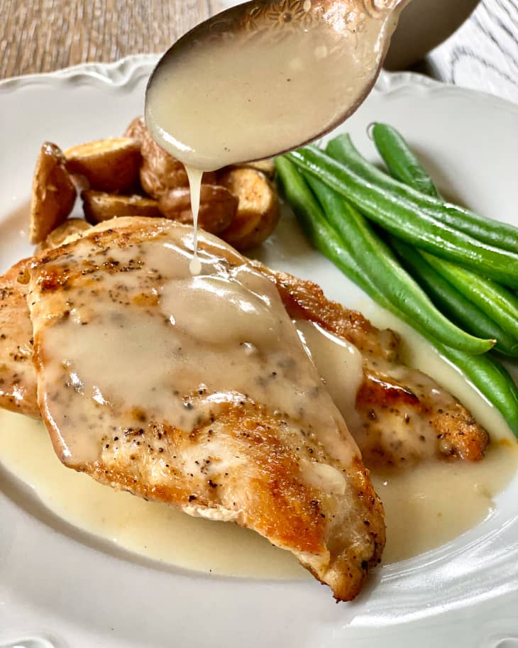 Veloute (a rich white sauce made with chicken, veal, pork, or fish stock, thickened with cream and egg yolks) being spooned over chicken cutlets, which are next to green beans, on a white plate.