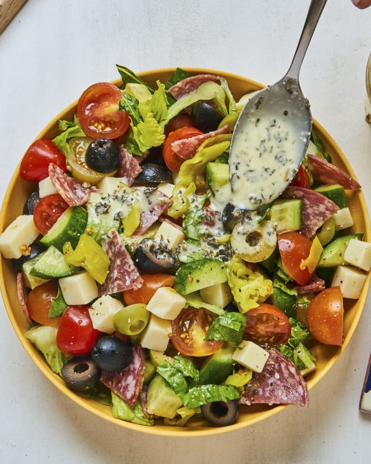 An Italian style salad with cucumbers, olives, tomatoes, cheese and cured meats, tossed in a creamy italian dressing, with a spoon with dressing sitting on the top of the salad.