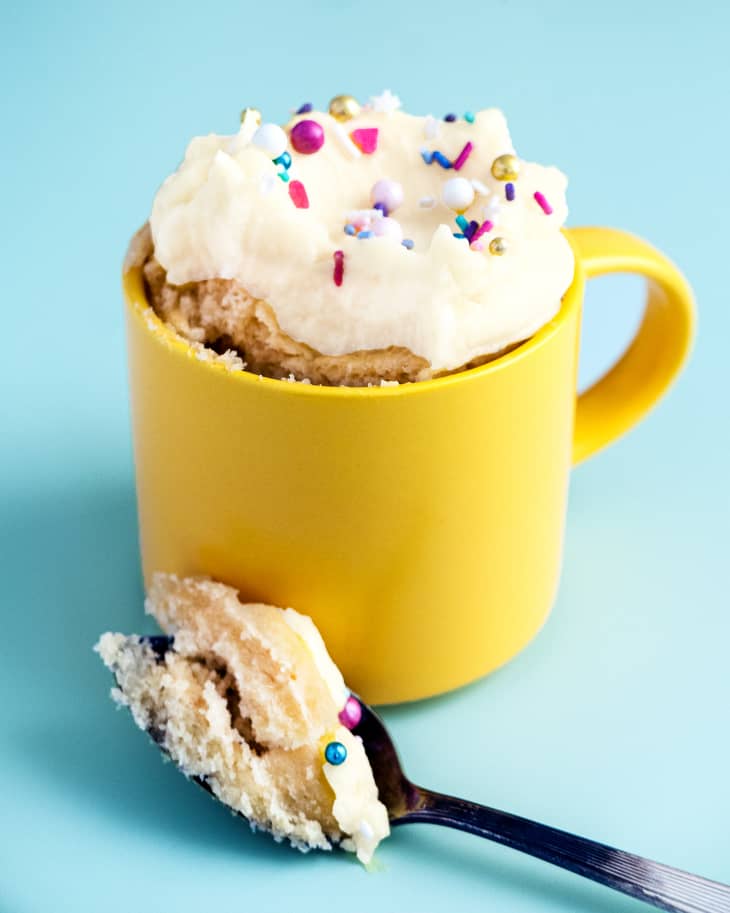 Vanilla cake baked in a yellow mug, with whipped cream and colored sprinkles on top. There is a spoon in front of the mug with a piece of the cake on it.