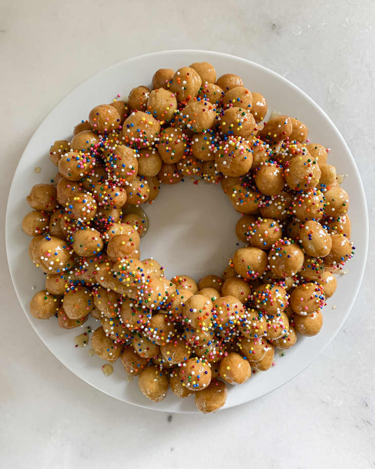 Struffoli, (Italian Honey Balls - very small crunchy dough balls mixed with honey and decorated with rainbow sprinkles) laid out in a donut shape on a circular white plate.