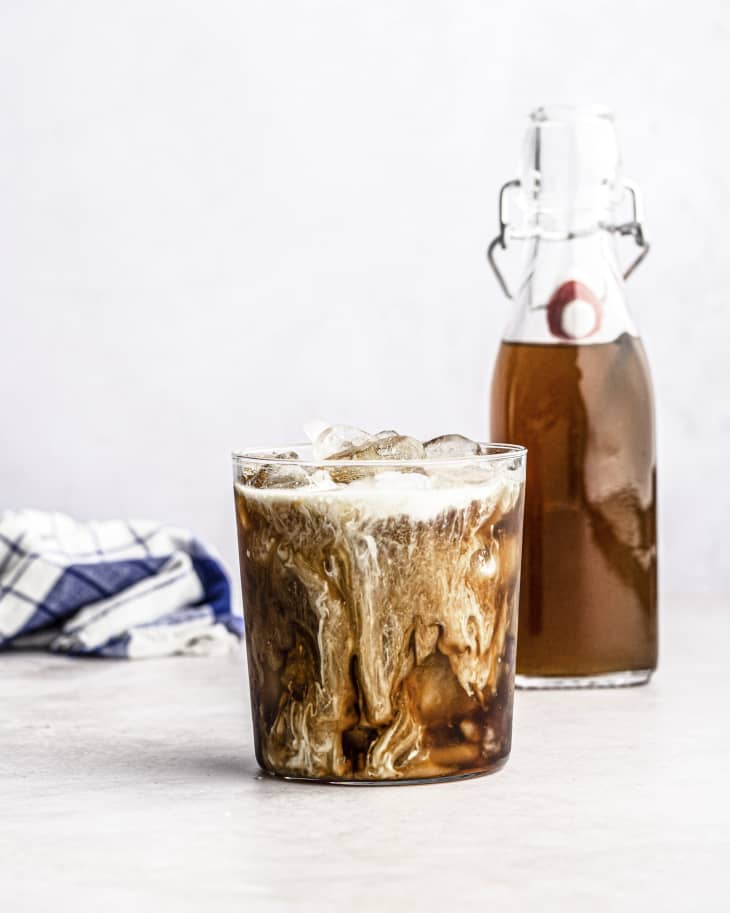 Vanilla syrup in a clear glass bottle with a plastic white stopper, with a stack of white and blue designed linen napkins next to it. A glass cup with coffee, syrup and cream barely mixed together so it appears marbleized, sits in front of it