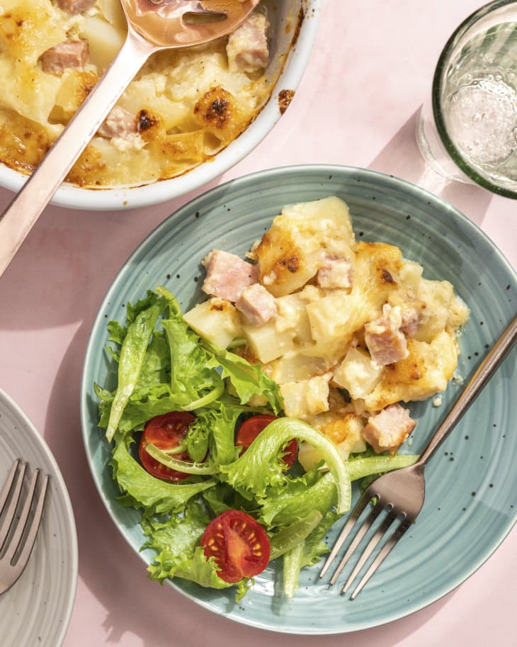 a blue plate with a scoop of ham and potato casserole, a green salad with tomatoes and a fork, with the ham and potato casserole dish next to it